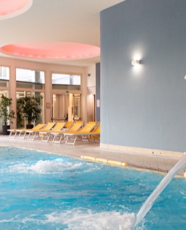 Interno di una spa con piscina azzurra, sdraio gialle e soffitto illuminato da luci rosa. Ambiente rilassante e moderno.