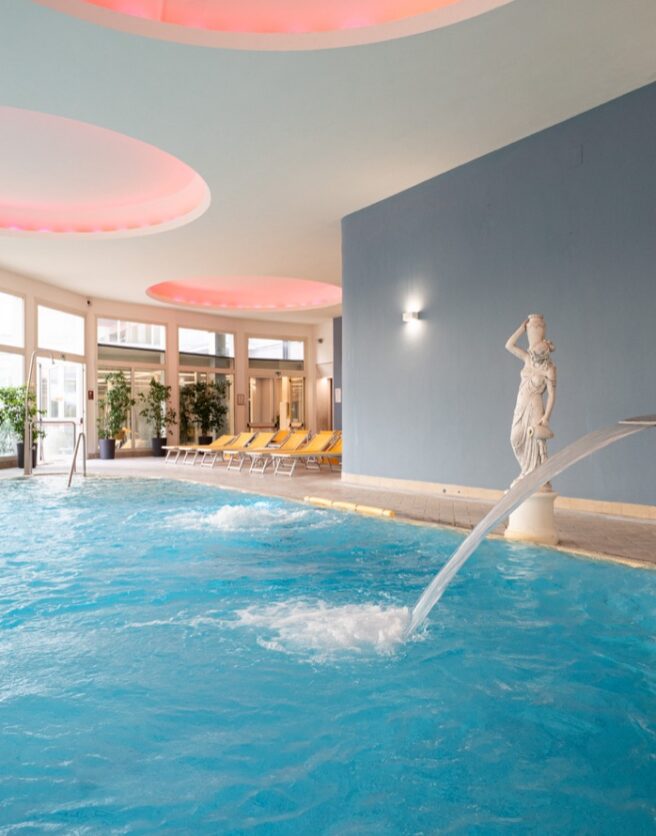 Piscina interna con statue e piante, soffitto rotondo illuminato in rosa, lettini a sdraio a bordo piscina.