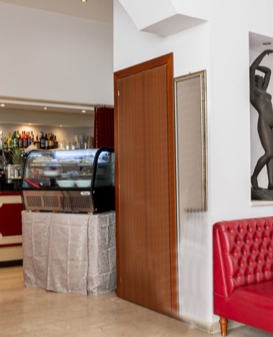 Interno di un caffè con una vetrina di dolci, una porta marrone e un divano rosso. Un'opera d'arte è appesa al muro.