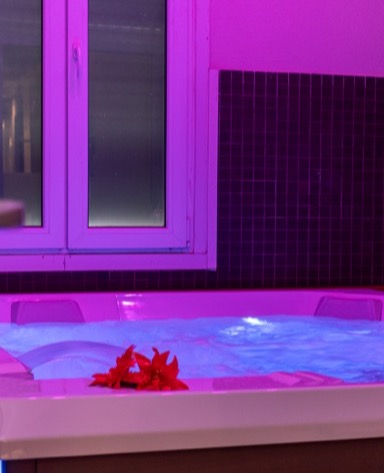 Vasca idromassaggio con acqua blu e fiori rossi, illuminata da luci viola, accanto a una finestra.