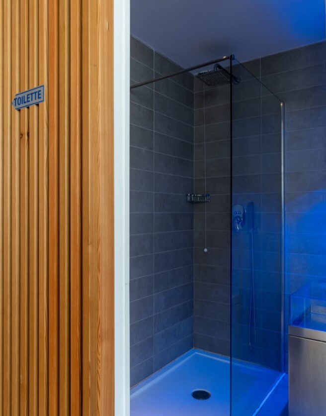 Area bagno con parete in legno, doccia a vetri e illuminazione blu, porta con segnale "TOILETTE".