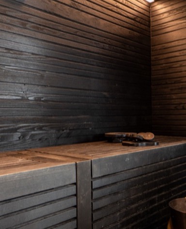 Parete nera con finitura a listelli e piano in legno chiaro, con alcuni oggetti metallici sopra.