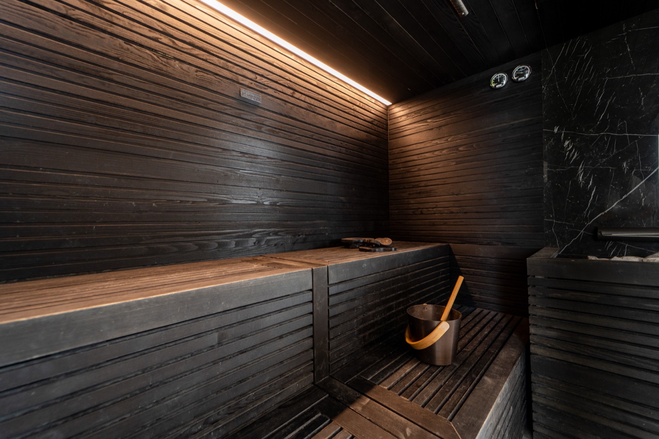 A modern sauna featuring dark wooden benches, a bucket with a wooden ladle, and minimalist black wall accents.