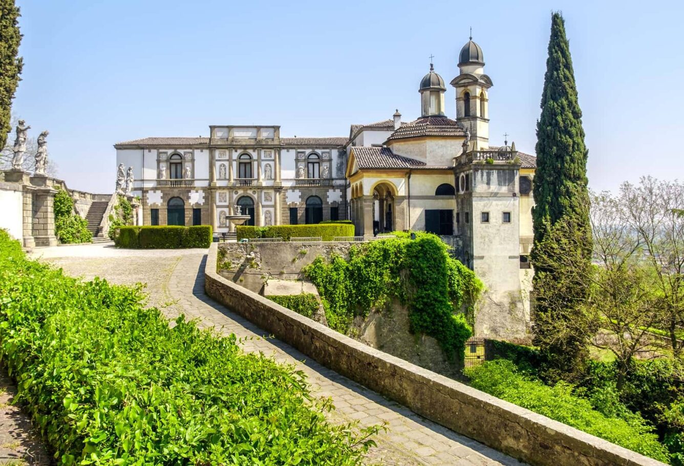 Edificio storico con elementi architettonici distintivi, circondato da giardini e alberi in una giornata soleggiata.